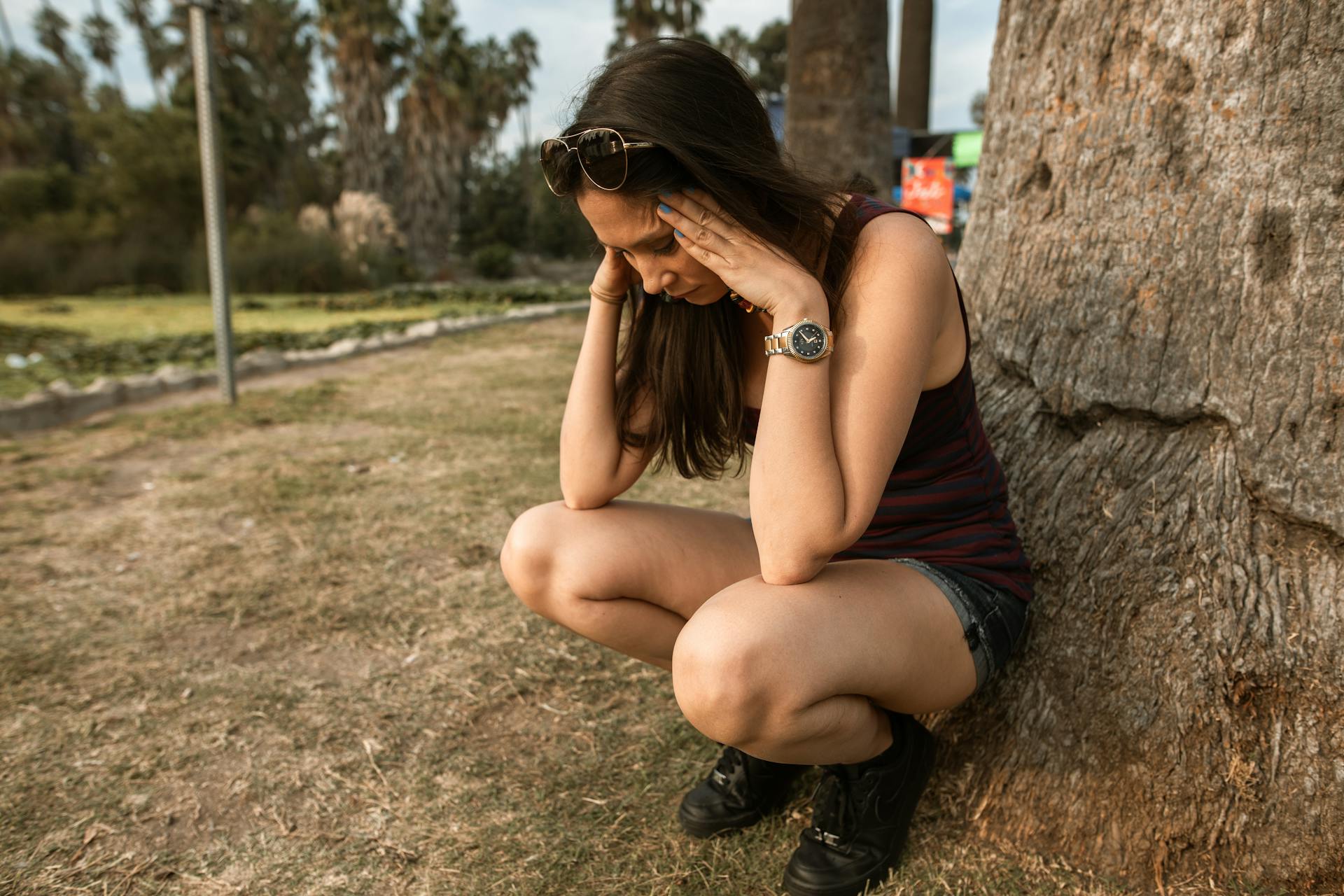 anxious woman crouches by a tree clutching her head